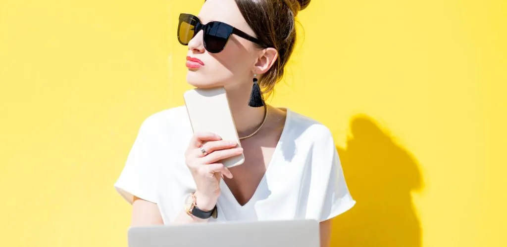 Woman deciding which CWS Israel pricing package she should purchase. She is wearing sunglasses and has her phone and laptop and is leaning against a bright yellow wall.